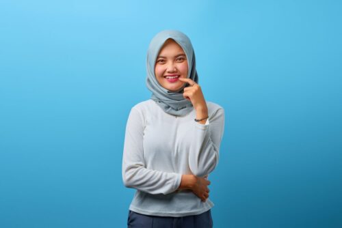 Cheerful teenage girl wearing hijab, posing in front of a bright blue backdrop.